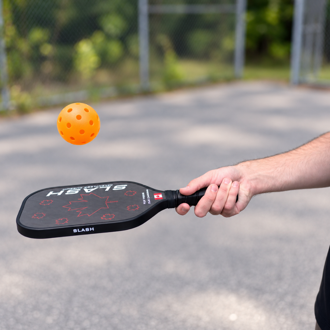 Orange pickleball in the air above a Slash paddle held by a player on an outdoor court